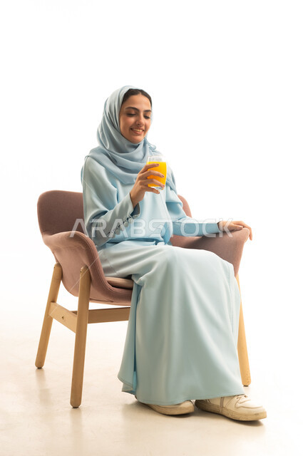 الاستمتاع بتناول المشروبات المفيدة اللذيذة، محاولة التخفيف من حرارة الصيف بالمشروب المنعش، بورترية لإمراة عربية سعودية خليجية محجبة ترتدي العباءة تجلس على كرسي تستمع بشرب العصير الطبيعي البارد، خلفية بيضاء