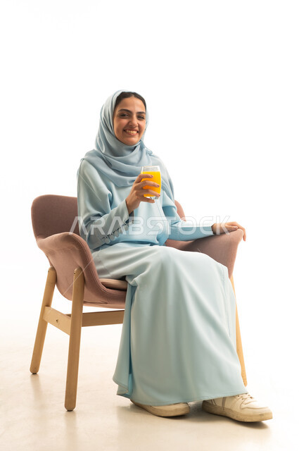 Trying to alleviate the summer heat with a refreshing drink, enjoying delicious and beneficial beverages, a portrait of a Saudi Arabian Gulf woman wearing a hijab and an abaya sitting on a chair enjoying a cold natural juice, with a white background.