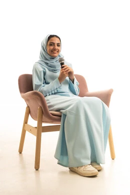 Performing theatrical shows, artistic talents in the world of music, musical theater art, practicing the hobby of singing for fun and entertainment, a portrait of an Arab Gulf Saudi woman wearing an abaya and hijab sitting on a chair holding a microphone in her hands, with a white background.