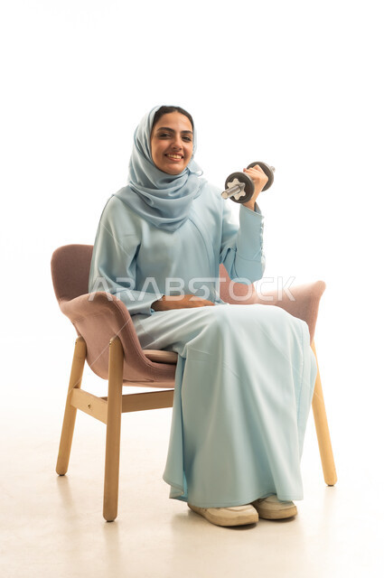ممارسة التمارين الرياضية ورفع الاثقال مع الدمبل، بورتريه لشابة عربية خليجية سعودية محجبة مبتسمة ترتدي العباءة تجلس على الكرسي المريح تحمل بيدها الأوزان الحديدية الثقيلة، المحافظة على الصحة واللياقة الجسدية والرشاقة البدنية، خلفية بيضاء