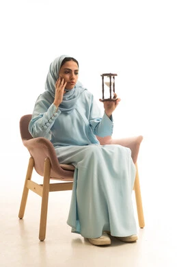 الجلوس بتعابير الملل والانتظار والسؤال, إدارة الأعمال وتنظيم الجدول الزمني, القيام بمراقبة توقيت الرمل, إدراك قيمة اللحظات الثمينة, بورتريه لشابة عربية خليجية سعودية ترتدي العباءة والحجاب تجلس على كرسي تمسك بيدها الساعة الرملية الزجاجية (البنكام), خلفية بيضاء