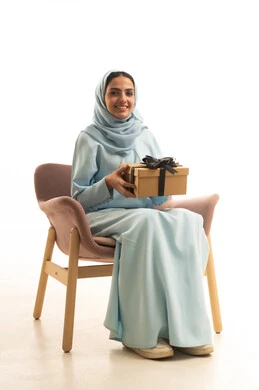 تقديم الهدايا في المناسبات السعيدة، إيماءات السعادة والسرور، الاحتفال بأعياد الميلاد, التعبير عن المحبة والتقدير, بورتريه لامرأة عربية خليجية سعودية محجبة ترتدي العباءة تجلس على كرسي تمسك بيديها علبة هدية, خلفية بيضاء