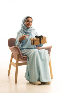 إيماءات السعادة والسرور، الاحتفال بأعياد الميلاد, التعبير عن المحبة والتقدير, تقديم الهدايا في المناسبات السعيدة, بورتريه لامرأة عربية خليجية سعودية محجبة ترتدي العباءة تجلس على كرسي تمسك بيديها علبة هدية و تشير اليها, خلفية بيضاء