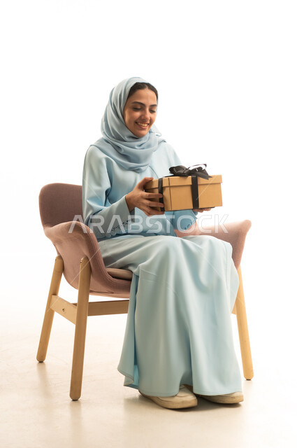 الاحتفال بأعياد الميلاد, التعبير عن المحبة والتقدير, تقديم الهدايا في المناسبات السعيدة, بورتريه لامرأة عربية خليجية سعودية محجبة ترتدي العباءة تجلس على كرسي تمسك بيديها علبة هدية,إيماءات السعادة والسرور, خلفية بيضاء