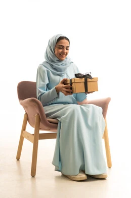Celebrating birthdays, expressing love and appreciation, giving gifts on happy occasions, a portrait of a Saudi Arabian Gulf woman wearing a hijab and an abaya sitting on a chair holding a gift box in her hands, gestures of happiness and joy, white background.