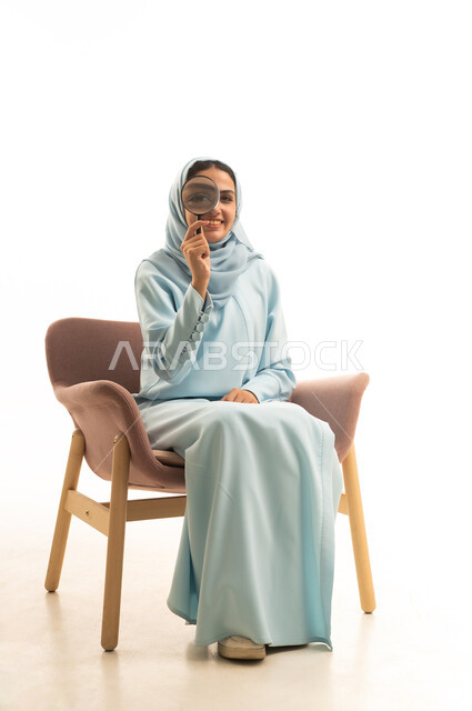 Scrutiny and focus using optical tools, the concept of image clarification, gestures of joy and happiness, examination and close vision, gestures indicating happiness and joy, a portrait of a Saudi Arabian Gulf woman wearing a hijab and an abaya sitting on a chair looking through a magnifying glass, white background.
