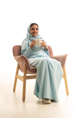 قضاء وقت ممتع و هادئ، الاستمتاع بتناول المشروب الساخن الشهي، بورتريه لامراه عربيه خليجيه سعودية محجبة ترتدي العباءه تجلس على كرسي تقوم بتناول كوب من الشاي , تعابيرالسعاده والسرور، خلفية بيضاء