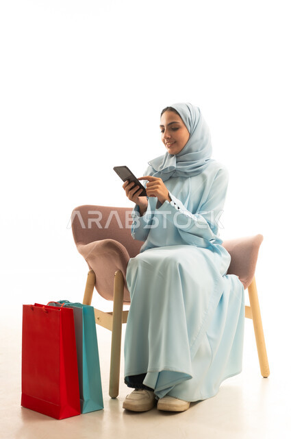 Mobile electronic marketing, ordering products and shopping online, seasonal offers and discounts, a portrait of a Gulf Arab Saudi woman wearing a hijab and an abaya holding a mobile phone sitting on a comfortable chair next to shopping bags, gestures of joy and happiness, white background.