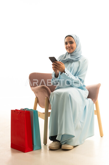 طلب المنتج والشراء عبر الانترنت, التسويق الإلكتروني عبر الجوال، عرض وتخفيض موسمي, بورتريه لإمرأة عربيه خليجية سعوديه محجبة ترتدي العباءة  تحمل بيدها الهاتف المحمول تجلس على كرسي مريح بجانب أكياس التسوق, إيماءات الفرح والسرور, خلفية بيضاء