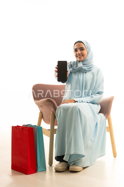 Shopping and purchasing supplies, using modern technological devices, a portrait of a Saudi Gulf Arab woman wearing a hijab and abaya sitting on a chair next to shopping bags holding a mobile phone with a blank black screen, white background.