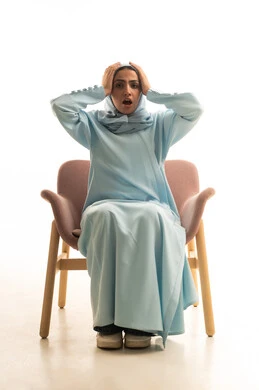 Surprise and astonishment at something, amazement and disbelief, loss and deprivation of something specific, gestures of wonder and astonishment, a portrait of an Arab Gulf Saudi woman wearing an abaya and hijab sitting on a chair raising her hands to her head with expressions of shock and astonishment, white background.