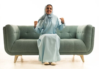 الحماس والشغف بمتابعة رياضة كرة القدم، مشاركة النساء السعوديات بالأنشطة الرياضية الترفيهية, بورتريه لامراة عربية خليجية سعودية ترتدي العباءة والحجاب تجلس على الاريكة  ترفع يدها بإيماءات السرور لفوز وانتصار الفريق المفضل, خلفية بيضاء