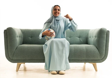 مشاركة النساء السعوديات بالأنشطة الرياضية الترفيهية, الحماس والشغف بمتابعة رياضة كرة القدم, بورتريه لامراة عربية خليجية سعودية ترتدي العباءة والحجاب تجلس على الاريكة تمسك كره القدم و ترفع يدها بإيماءات السرور لفوز وانتصار الفريق المفضل, خلفية بيضاء