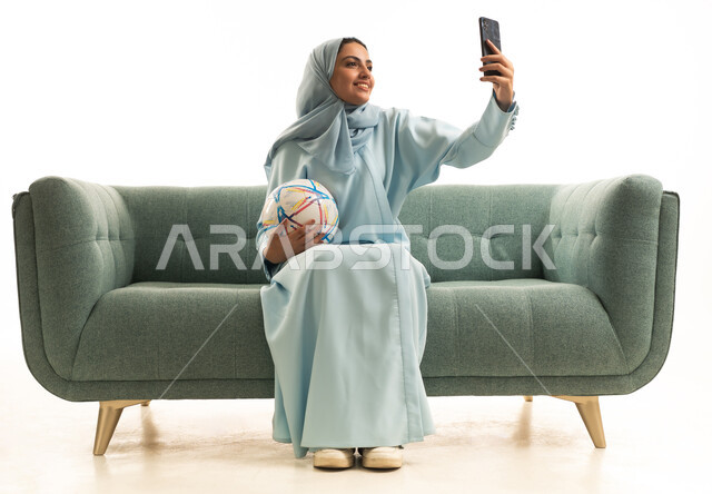 Taking selfies with a mobile phone, the passion for football, matches and sports, a portrait of a Saudi Gulf Arab woman wearing a hijab and an abaya sitting on a sofa holding a football and a mobile phone, white background.