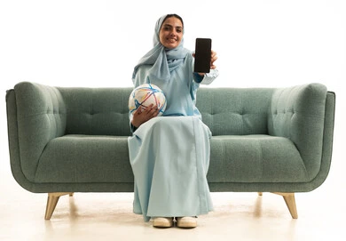 عرض شاشة سوداء عبر الهاتف المحمول ، الشغف برياضة كرة القدم ، المباريات والألعاب الرياضية، بورتريه لامراه عربية خليجيه سعودية محجبة ترتدي العباءة تجلس على الاريكة تمسك كرة قدم, خلفية بيضاء