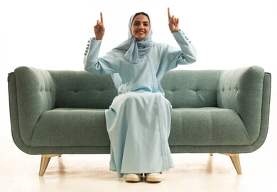 Identifying a specific location with gestures of cheerfulness, looking at something with expressions of joy, signals and hand movements indicating this place (here), a portrait of a smiling, veiled Saudi Arabian Gulf woman wearing an abaya sitting on a sofa, raising her fingers and pointing upwards, with a white background.