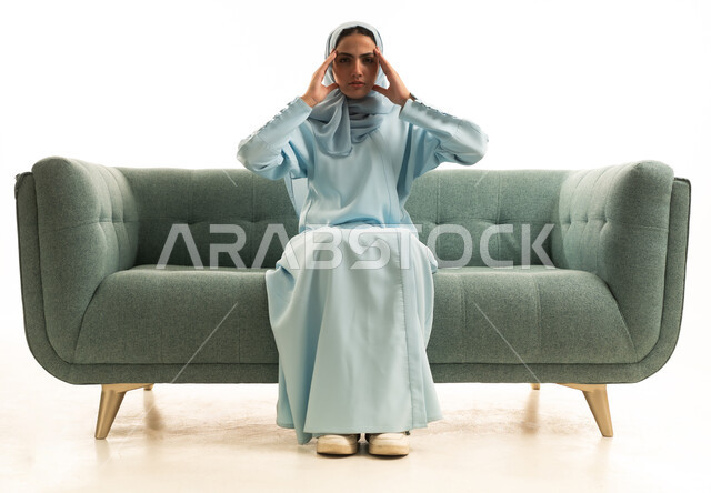 The feeling of headache and migraine, the need for calm and rest, suffering from migraine pain, a portrait of a Saudi Arabian Gulf woman wearing a hijab and an abaya sitting on the sofa with her hands on her head showing signs of fatigue and exhaustion, white background.