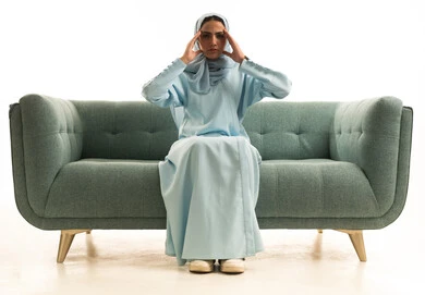 الإحساس بألم الرأس والصداع، الحاجة للهدوء والراحة، المعاناة من آلام الشقيقة، بورتريه لإمرأة عربية خليجية سعودية محجبة ترتدي العباءة تجلس على الاريكة تضع يديها على رأسها بايماءات التعب والارهاق، خلفية بيضاء