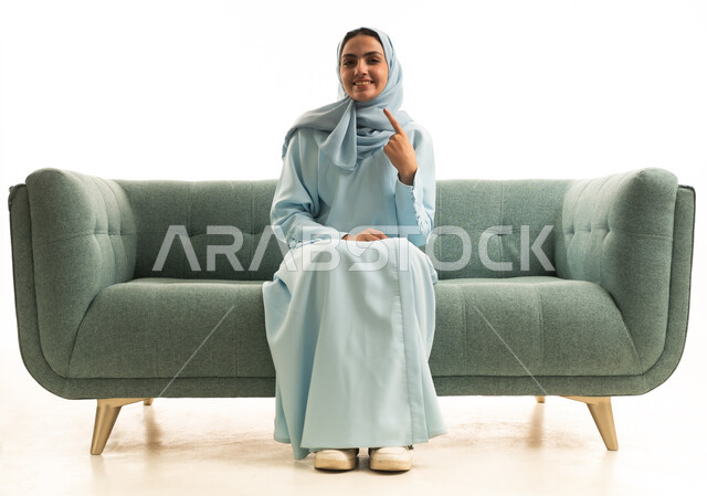 إبتسامة هوليود باستخدام قشور فينير من الخزف، بورتريه لامراة عربية خليجية سعودية مبتسمة ترتدي العباءة والحجاب تجلس على الاريكة تنظر إلى الكاميرا وتشير بإصبع السبابة على أسنانها، العناية والاهتمام بالصحة الفموية، خلفية بيضاء