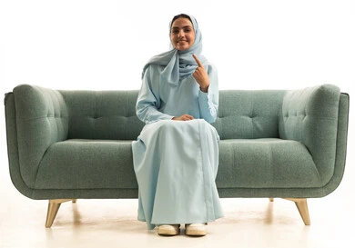 إبتسامة هوليود باستخدام قشور فينير من الخزف، بورتريه لامراة عربية خليجية سعودية مبتسمة ترتدي العباءة والحجاب تجلس على الاريكة تنظر إلى الكاميرا وتشير بإصبع السبابة على أسنانها، العناية والاهتمام بالصحة الفموية، خلفية بيضاء