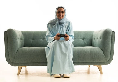 ايماءات التفاعل و الاندماج، قضاء أوقات الفراغ باللعب بألعاب البلايستيشن الترفيهية ، تقنيات حديثة مسلية, بورتريه لإمرأة عربية سعودية خليجية تجلس على  الاريكة وتحمل بيدها عصا التحكم و تلعب بألعاب الفيديو ، خلفية بيضاء