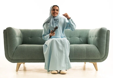 إيماءات وتعابير وجه ويدين تدل على الفوز و الانتصار, قضاء أوقات الفراغ باللعب بألعاب البلايستيشن الترفيهية ، تقنيات حديثة مسلية, بورتريه لإمرأة عربية سعودية خليجية تجلس على  الاريكة وتحمل بيدها عصا التحكم و تلعب بألعاب الفيديو ، خلفية بيضاء