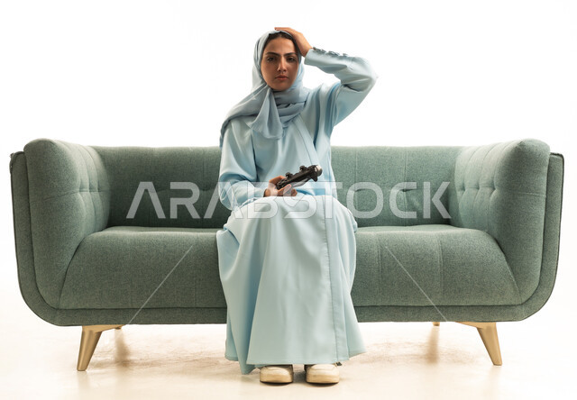 اللعب بألعاب الفيديو الترفيهية، تعابير الحزن والخسارة, استخدام الأجهزة التكنولوجية والتقنيات الحديثة،  بورتريه لامراة عربية خليجية سعودية محجبة ترتدي العباءة تجلس على الاريكة تمسك عصا التحكم تضع يدها على رأسها بإيماءات الحسرة، خلفية بيضاء