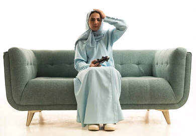 اللعب بألعاب الفيديو الترفيهية، تعابير الحزن والخسارة, استخدام الأجهزة التكنولوجية والتقنيات الحديثة،  بورتريه لامراة عربية خليجية سعودية محجبة ترتدي العباءة تجلس على الاريكة تمسك عصا التحكم تضع يدها على رأسها بإيماءات الحسرة، خلفية بيضاء