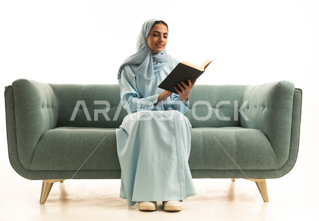التعلم وزيادة الوعي عن طريق قراءة الكتب، ممارسة هواية المطالعة, مفهوم الشغف بالقراءة, بورتريه لامراة عربية خليجية سعودية محجبة ترتدي العباءة تجلس على  الاريكة تقوم بقراءه كتاب, كتب وروايات هادفه ومسليه، خلفيه بيضاء