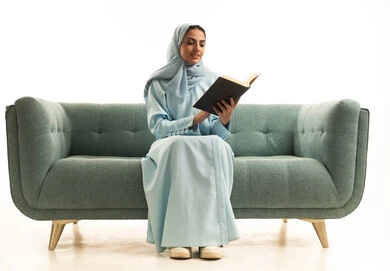 Learning and increasing awareness through reading books, practicing the hobby of reading, the concept of passion for reading, a portrait of a Saudi Gulf Arab woman wearing a hijab and an abaya sitting on a sofa reading a book, purposeful and entertaining books and novels, white background.