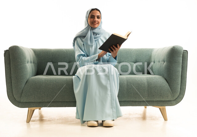 Developing knowledge and culture through reading books, practicing the hobby of reading, the concept of passion for reading, a portrait of a Saudi Gulf Arab woman wearing a hijab and an abaya sitting on a sofa reading a book, purposeful and entertaining books and novels, white background.