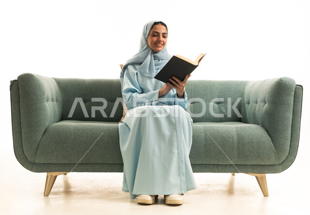 اكتساب المعرفة والتثقيف عبر قراءة الكتب، ممارسة هواية المطالعة, مفهوم الشغف بالقراءة, بورتريه لامراة عربية خليجية سعودية محجبة ترتدي العباءة تجلس على  الاريكة تقوم بقراءه كتاب, كتب وروايات هادفه ومسليه، خلفيه بيضاء
