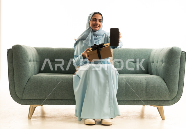 عرض شاشة سوداء عبر الجوال، الاحتفال بأعياد الميلاد, التعبير عن المحبة والتقدير,تقديم الهدايا في المناسبات السعيدة, بورتريه لإمراة عربية خليجية سعودية ترتدي الحجاب والعباءة تجلس على الاريكة تمسك بيديها علبة هداية و الهاتف المحمول, خلفية بيضاء