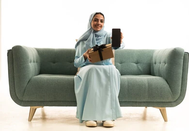 عرض شاشة سوداء عبر الجوال، الاحتفال بأعياد الميلاد, التعبير عن المحبة والتقدير,تقديم الهدايا في المناسبات السعيدة, بورتريه لإمراة عربية خليجية سعودية ترتدي الحجاب والعباءة تجلس على الاريكة تمسك بيديها علبة هداية و الهاتف المحمول, خلفية بيضاء