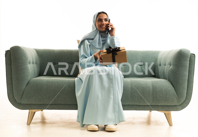 اجراء مكالمة هاتفية عبر الجوال، الاحتفال بأعياد الميلاد, التعبير عن المحبة والتقدير,تقديم الهدايا في المناسبات السعيدة, بورتريه لإمراة عربية خليجية سعودية ترتدي الحجاب والعباءة تجلس على الاريكة تمسك بيديها علبة هداية و الهاتف المحمول, خلفية بيضاء