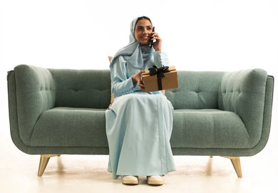 Making a phone call via mobile, celebrating birthdays, expressing love and appreciation, giving gifts on happy occasions, a portrait of a Gulf Arab Saudi woman wearing a hijab and abaya sitting on a sofa holding a gift box and a mobile phone, white background.