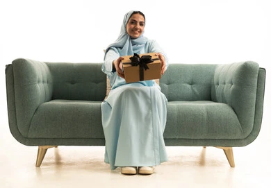 الاحتفال بأعياد الميلاد, التعبير عن المحبة والتقدير,تقديم الهدايا في المناسبات السعيدة, بورتريه لإمراة عربية خليجية سعودية ترتدي الحجاب والعباءة تجلس على الاريكة تمسك بيديها علبة هداية, خلفية بيضاء