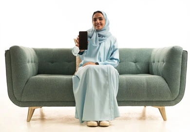 Electronic marketing using technology and tech, showcasing mobile applications, a portrait of a Saudi Gulf Arab woman wearing a hijab and abaya sitting on a sofa holding a mobile phone with a blank black screen, white background.