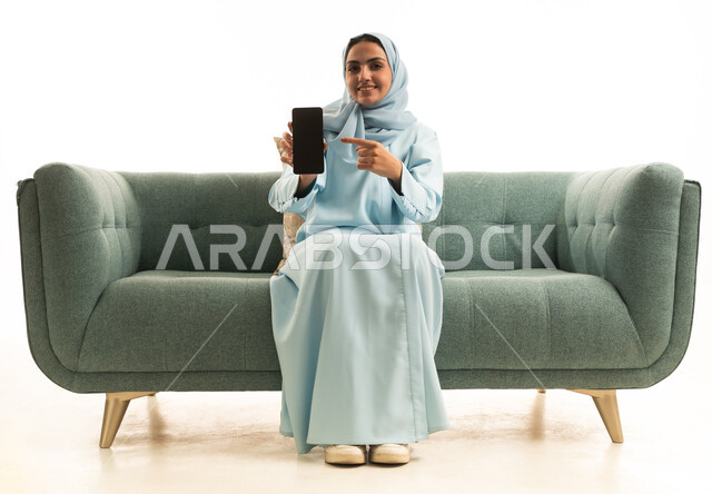 عرض تطبيقات الهاتف المحمول، التسويق الالكتروني باستخدام التقنية والتكنولوجيا، بورتريه لإمراة عربية خليجية سعودية ترتدي الحجاب والعباءة تجلس على الاريكة تحمل بيدها جوال بشاشة سوداء فارغة و تشير اليها، خلفية بيضاء