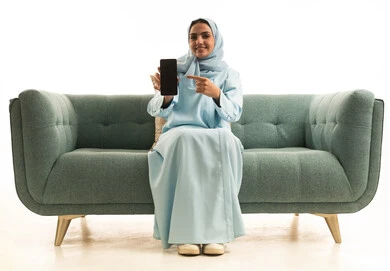 عرض تطبيقات الهاتف المحمول، التسويق الالكتروني باستخدام التقنية والتكنولوجيا، بورتريه لإمراة عربية خليجية سعودية ترتدي الحجاب والعباءة تجلس على الاريكة تحمل بيدها جوال بشاشة سوداء فارغة و تشير اليها، خلفية بيضاء