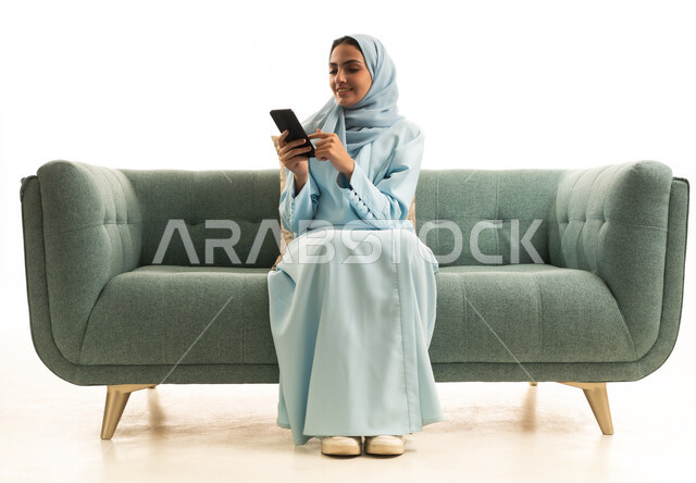 Browsing applications and social media platforms, exchanging conversations and news with family and friends, a portrait of a Saudi Gulf Arab woman wearing a hijab and an abaya sitting on a sofa holding a mobile phone, using modern advanced devices and technologies, white background.