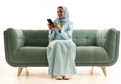 تصفح التطبيقات وبرامج التواصل الاجتماعي، تبادل الأحاديث والأخبار مع الأهل والأصدقاء، بورتريه لامراة عربية خليجية سعودية محجبة ترتدي العباءة تجلس على الاريكة تمسك بيدها الهاتف المحمول، استخدام الأجهزة والتقنيات الحديثة المتطورة، خلفية بيضاء