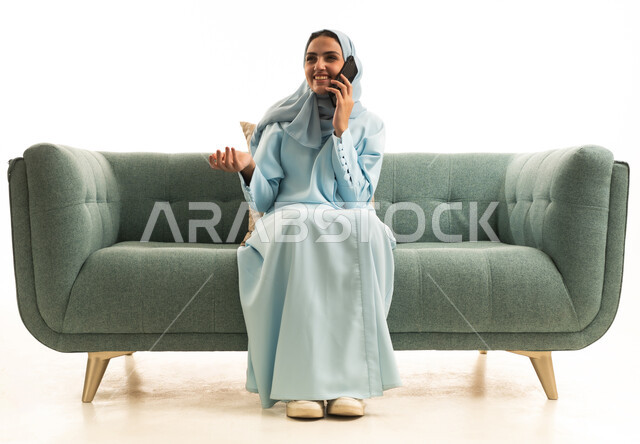 التواصل مع العائلة والأصدقاء, استخدام الأجهزة التقنية الحديثة في الحياة اليومية, مشاركة الأخبار والأحاديث الجيدة, بورتريه لامرأة عربية خليجية سعودية محجبة ترتدي العباءة تجلس على الاريكة تمسك بيدها الهاتف المحمول, إجراء اتصال هاتفي، خلفية بيضاء