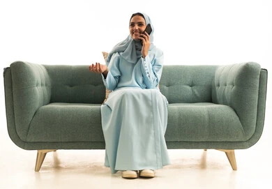 Communicating with family and friends, using modern technological devices in daily life, sharing news and good conversations, a portrait of a Saudi Arabian Gulf woman wearing a hijab and an abaya sitting on a sofa holding a mobile phone, making a phone call, white background.