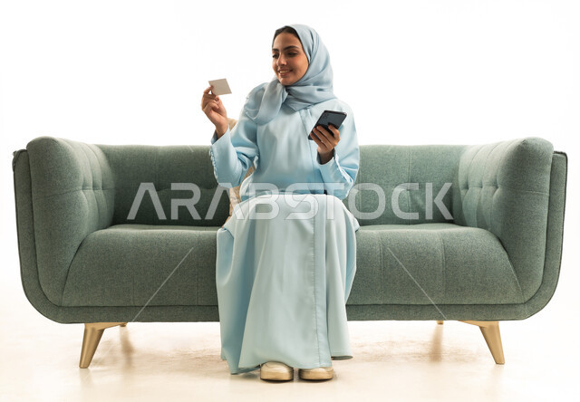 استخدام جهاز تقني حديث متطور، الدفع عبر الانترنت، إمكانية الشراء والدفع من خلال برامج وتطبيقات الجوال،بورترية لإمرأة عربية خليجية سعودية مبتسمة ترتدي الحجاب تجلس على الاريكة تحمل الهاتف المحمول وبطاقة الإئتمان البنكية، خلفية بيضاء
