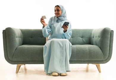 استخدام جهاز تقني حديث متطور، الدفع عبر الانترنت، إمكانية الشراء والدفع من خلال برامج وتطبيقات الجوال،بورترية لإمرأة عربية خليجية سعودية مبتسمة ترتدي الحجاب تجلس على الاريكة تحمل الهاتف المحمول وبطاقة الإئتمان البنكية، خلفية بيضاء