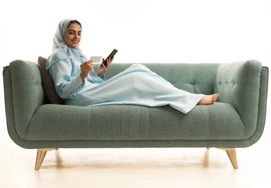 The possibility of paying bills via credit card, purchasing and paying through mobile apps and programs, a portrait of a smiling Arab Gulf Saudi woman wearing a hijab sitting on a sofa holding a mobile phone and a credit card, white background.