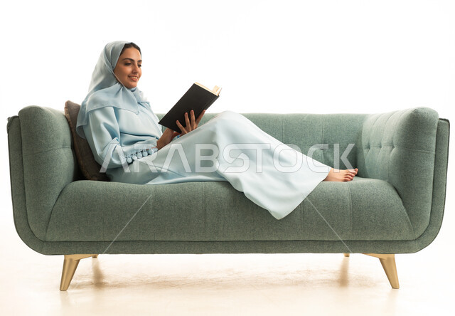كتب وروايات هادفه ومسليه، مفهوم الشغف بالقراءة، ممارسة هواية المطالعة, بورتريه لامراة عربية خليجية سعودية محجبة ترتدي العباءة تجلس على  الاريكة تقوم بقراءه كتاب، التعلم والتثقيف من خلال قراءه الكتب الهادفه، خلفيه بيضاء