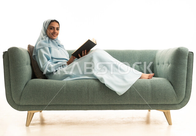 مفهوم الشغف بالقراءة، ممارسة هواية المطالعة, بورتريه لامراة عربية خليجية سعودية محجبة ترتدي العباءة تجلس على  الاريكة تقوم بقراءه كتاب, كتب وروايات هادفه ومسليه ، التعلم والتثقيف من خلال قراءه الكتب الهادفه، خلفيه بيضاء