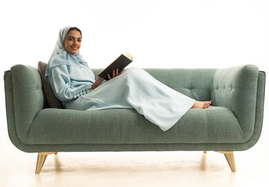 The concept of passion for reading, practicing the hobby of reading, a portrait of a Gulf Arab Saudi woman wearing a hijab and an abaya sitting on a sofa reading a book, purposeful and entertaining books and novels, learning and educating through reading purposeful books, white background.